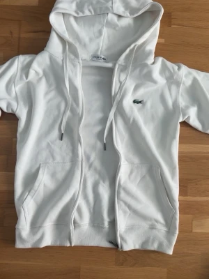 Vit hoodie från Lacoste med dragkedja - Snygg vit hoodie från Lacoste i regular fit med dragkedja framtill, två fickor och klassisk huva med snören. Ikoniska gröna krokodilloggan på bröstet. Perfekt för en clean och sportig stil.