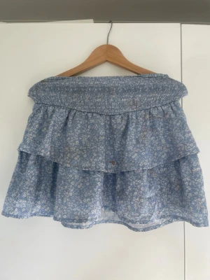 Blommig blå volangkjol - Söt kort blå kjol med små vita blommor och volangdetaljer. Kjolen har en bred smockresår i midjan för en bekväm passform och är perfekt för varma dagar. Lätt och luftig känsla, passar dig som gillar romantisk och avslappnad stil. Kjolen har orangea fläckar troligen från foundation vilket går att tvätta bort. 