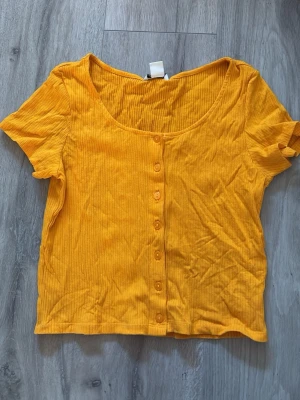 Gul  topp från Monki - Superfin gul ribbad topp från Monki med korta ärmar och knappar framtill. Toppen har en rundad halsringning och är i mjukt, stretchigt material som sitter snyggt på kroppen. Perfekt att styla med jeans eller kjol för en färgglad look. Knappt använd