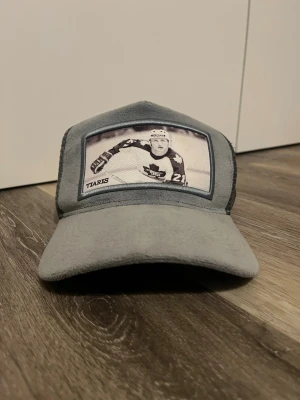Grå ”Börje Salming” keps från ”Brand of Tiaris” med tryck - Cool grå keps från TIARIS med mörkgrå mesh på sidorna och ett stort hockeymotiv framtill. Kepsen har böjd skärm och justerbar baksida. Perfekt för dig som gillar sport och vill sticka ut med en unik accessoar. Kepsen är ljusblå/grå.  Pris kan absolut diskuteras.
