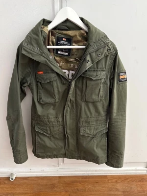 Grön vindjacka från Superdry - Stilren militärgrön vindjacka från Superdry med fyra stora fickor framtill, huva och patchdetaljer på bröstet och ärmen. Jackan har dragkedja och tryckknappar samt camouflagemönstrat innerfoder. Perfekt för en urban streetlook. Strl:S✅ Pris kan diskuteras!💸 Hör av dig om du har några funderingar!👍