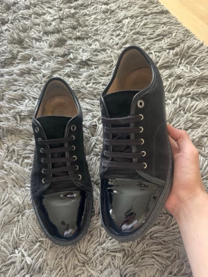 Svarta sneakers från Lanvin - Snygga svarta sneakers från Lanvin med en mix av mocka och lackdetaljer på tån. Skorna har svarta skosnören, metallöljetter och är tillverkade i Portugal. Insidan är fodrad i beige läder för extra komfort. Låda och dustbag ingår