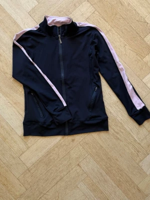 Svart träningsjacka med rosa detaljer - Svart träningsjacka med hel dragkedja och ljusrosa ränder längs ärmarna. Jackan har hög krage, två fickor med dragkedja och är tillverkad i ett stretchigt, sportigt material. Perfekt för träning eller som snyggt lager till vardags.