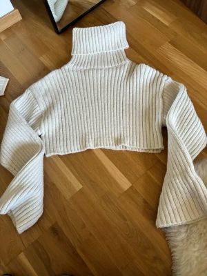 Vit cropped polotröja från Zara - Säljer en vit ribbstickad cropped polotröja från Zara. Tröjan har långa vida ärmar och hög polokrage, perfekt för lager-på-lager. Mjuk och skön stickad kvalitet som ger en trendig look.