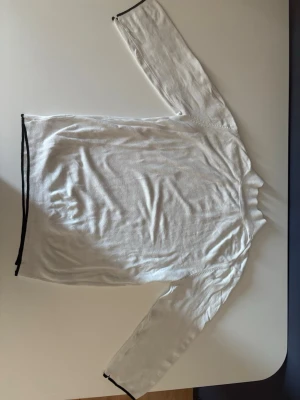 Vit långärmad tröja från Zara - Vit långärmad tröja i tunnare material med svarta kanter vid ärmslut och nederkant. Enkel och stilren design, perfekt för lager-på-lager eller som basplagg. Rund halsringning och rak passform.