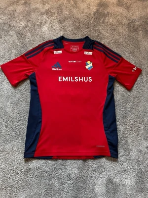 Östers IF Röd och marinblå hemmatröja 2024/25 Adidas - Snygg röd och marinblå fotbollströja från Adidas med Östers IF klubbmärke och flera sponsorer tryckta på bröstet och ryggen. Tröjan har korta ärmar, rund hals och är tillverkad i ett lätt och ventilerande funktionsmaterial. Perfekt för träning eller match, men också everyday wear.