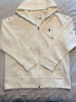 Vit hoodie från Polo Ralph Lauren - Snygg vit hoodie från Polo Ralph Lauren med dragkedja framtill och klassisk huva. Broderad logga i svart på bröstet och två fickor på sidorna. Perfekt för en clean och stilren look. Tillverkad i mjukt bomullsmaterial. I storlek M.                              Vid några funderingar är det bara att höra av sig☺️