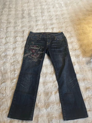 Mörkblå jeans med brodyr från Ed Hardy - Snygga mörkblå jeans från Ed Hardy med coolt broderat motiv på ena låret. Klassisk femficksmodell med raka ben och slitningar för en trendig look. Jeansen har detaljer i gult på bakfickan och är tillverkade i bomullsdenim.säljer detta pågrund av att dom är för korta för mig