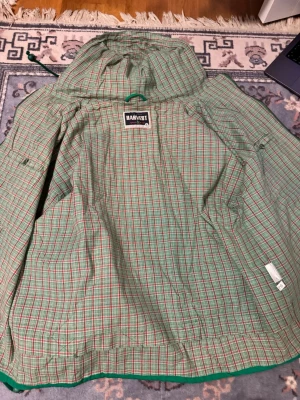 Grön regnponcho från Harvest - Grön regnponcho från Harvest med rutigt innerfoder i grönt, rött och vitt. Ponchon har huva med dragsko och är tillverkad i ett lätt material som skyddar mot regn. Perfekt för utomhusaktiviteter när vädret är ostadigt.