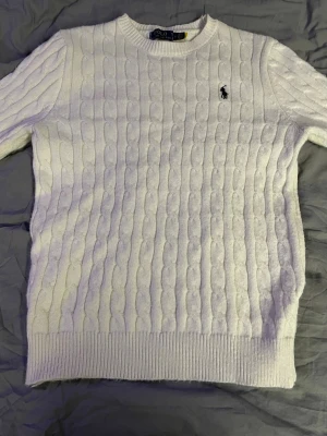 Vit kabelstickad tröja Polo Ralph Lauren - Vit kabelstickad tröja från Polo Ralph Lauren med rund halsringning och broderad logga på bröstet. Tröjan har ribbade muddar vid ärmslut och nederkant, perfekt för en clean och klassisk look.
