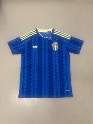 Sveriges vm tröja gyökeres - Blå fotbollströja från Adidas med Sveriges landslagsemblem och gula detaljer. Tröjan har mönstrade ränder, gula axelband och namnet 'GYÖKERES' på ryggen. Perfekt för dig som vill stötta Sverige på match eller träning.
