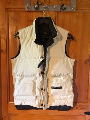 Vit dunväst från Canada goose - Stilren vit dunväst från Canada goose med svarta detaljer och hög krage. Västen har två stora fickor framtill, tryckknappar och en diskret logotyp på ena fickan. Perfekt för lager-på-lager och håller dig varm under kyliga dagar.