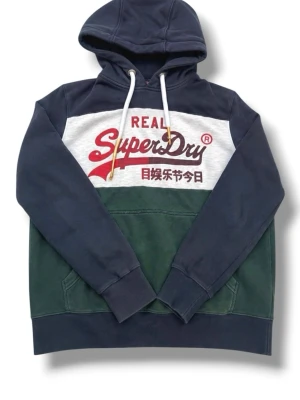 Superdry hoodie - Snygg hoodie från Superdry med färgblock i marinblått, vitt och mörkgrönt. Stor logga och japansk text framtill, klassisk känguruficka och justerbar huva med vita snören. Perfekt för en avslappnad streetstyle-look.