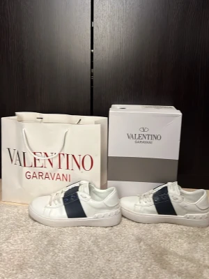 Valentino Garavani vita sneakers - Snygga vita sneakers från Valentino Garavani med en mörkblå detalj på sidan. Skorna har klassisk rund tå, platt sula och snörning. Tillverkade i skinn för en lyxig känsla och stilren look. Perfekta för dig som vill ha något exklusivt och trendigt. Mitt pris 1800 nypris 6000:-