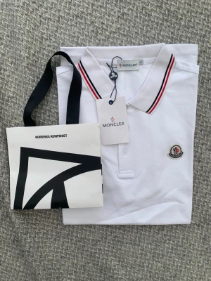 Vit pikétopp från Moncler - Vit pikétopp från Moncler med klassisk krage och knäppning framtill. Kragen har snygga ränder i svart och rött. Moncler-logga broderad på bröstet. Perfekt för en stilren och sportig look.