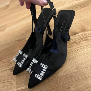 Svarta pumps med strass från Sole Sisters - Snygga svarta pumps med spetsig tå och hög klack från Sole Sisters. Skorna har en elegant rem runt hälen med spänne och är dekorerade med en stor strassdetalj framtill. Materialet är satinliknande och ger en lyxig känsla. Perfekt för dig som gillar att sticka ut. Två stenar har lossnat, men de är i gott skick och det syns nästan inte.