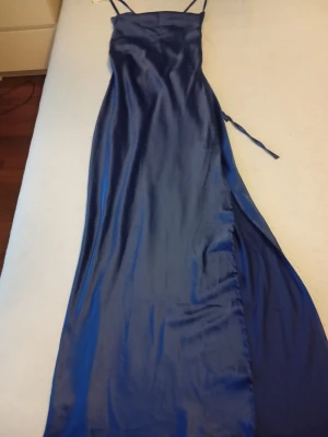 Blå långklänning med öppen rygg - Elegant blå långklänning i satin med smala axelband och snygg öppen rygg med snörning. Klänningen har en slits på sidan och ett glansigt material som ger en lyxig känsla. Perfekt för dig som vill sticka ut med en stilren look.