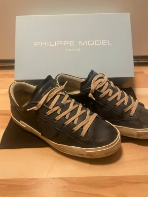 Philippe Model svarta skinnsneakers - Säljer ett par Philippe Model sneakers i svart skinn med beige snörning och detaljer. Klassisk låg modell med vit sula och diskret logga på sidan. Perfekta för dig som gillar stilrena och exklusiva sneakers.