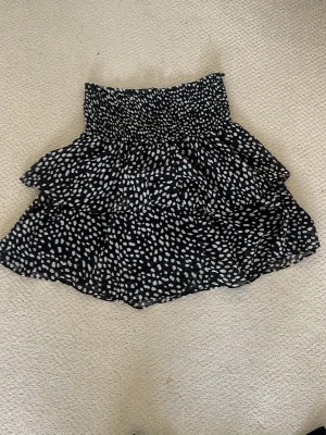 Svart vollang-kort-kjol med vita prickar - Säljer en svart kortkjol med vita prickar och volangdetaljer. Kjolen har en smockad midja som ger en bekväm och snygg passform. Storlek XS/S, men är mycket stretchig. Pris diskuteras gärna och skicka gärna om ni har frågor! Köpt på Chelsea!