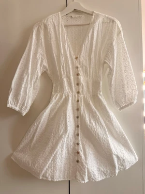 Vit kort klänning med broderi - Söt vit kortklänning med broderade detaljer och knappar framtill. Klänningen har v-ringning, trekvartsärm och markerad midja för en feminin siluett. Tillverkad i luftig bomull, perfekt för studenten. Klänningen är i XS men funkar utmärkt som S