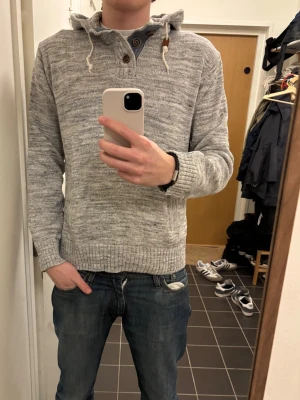 Gråmelerad hoodie med knappar - Säljer en gråmelerad hoodie med huva och dragsnören. Tröjan har ribbade muddar, knappslå med fyra knappar framtill och fickor på sidorna. Materialet är mjukt och stickat, perfekt för en avslappnad stil.