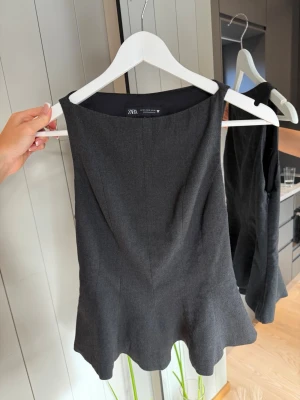 Mörkgrå peplum topp från Zara - Säljer en mörkgrå ärmlös topp från Zara med peplumkant. Toppen har en stilren, figurnära passform och bred halsringning. Perfekt att styla med både jeans och kjol för en trendig look.