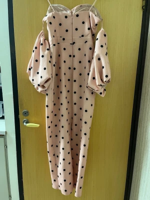 Prickig långklänning med puffärmar - Supersnygg långklänning i ljus beige med svarta prickar. Klänningen har puffiga offshoulder-ärmar, smala axelband och ett dekorativt bälte med strassdetalj i midjan. V-ringad upptill och figurnära passform. Perfekt för dig som vill sticka ut!