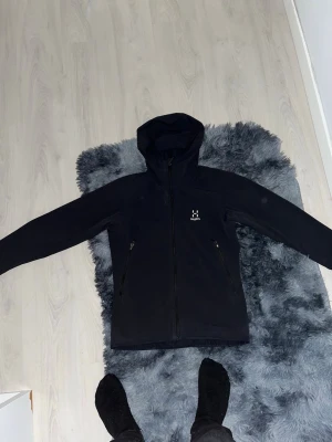 Svart vindjacka från Haglöfs - Snygg svart vindjacka från Haglöfs med huva och dragkedja framtill. Två praktiska sidofickor med dragkedja och diskret logga på bröstet. Jackan har en stilren design och är tillverkad i ett lätt och vindtätt material.