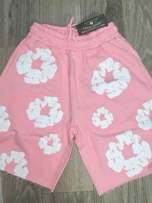 Rosa shorts med vita blommor - Säljer ett par rosa shorts från Denim Tears med vita blommönster. Shortsen har resårmidja med snörning och råa kanter nertill för en avslappnad look. Perfekta för varma dagar och ger en trendig vibe.