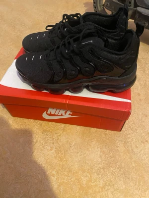 Nike Air Vapormax Plus svarta sneakers - Nike Air Vapormax Plus sneakers i helsvart med tydliga detaljer och synlig luftdämpning i sulan. Ovandelen är i textil och syntet med svarta snören och vita accenter på tån. Snygg, futuristisk design som sticker ut och passar perfekt till streetwear.
