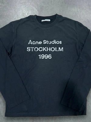 Acne Studios Svart långärmad - Cool svart långärmad topp från Acne Studios med vit text 'STOCKHOLM 1996' på bröstet. Klassisk rund halsringning och relaxed fit, perfekt att styla med jeans. Materialet är mjuk bomull som känns skönt mot huden.