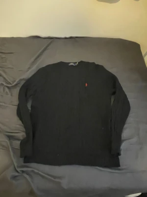 Svart kabelstickad tröja Polo Ralph Lauren - Kabelstickad svart tröja från Polo Ralph Lauren med den klassiska röda loggan broderad på bröstet. Tröjan har rund halsringning och långa ärmar. Perfekt för dig som gillar stilrena och tidlösa plagg.