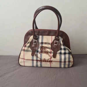 Rutig Burberry handväska med bruna detaljer - Snygg handväska från Burberry med klassiskt rutigt mönster i beige, brun, blått och röd. Väskan har bruna handtag och detaljer i krokopräglat skinn, samt broderad logga framtill. Praktisk dragkedja upptill(ena är trasig genom att den inte stänger nör men drar i den men eftersom det finns två funkar den fortfarande bra)och metallspännen på handtagen. Perfekt för dig som gillar ikonisk design. Jag köpte den på loppis, säljaren sa att den var äkta men jag vet inte om det stämmer
