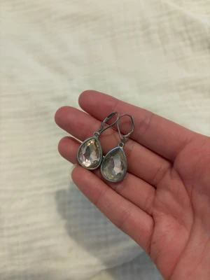 Silvriga örhängen med droppformad sten - Snygga örhängen i silverfärg med stora, droppformade klara stenar. Klassisk design med fasettslipad yta som fångar ljuset. Perfekta för dig som gillar stilrena accessoarer med lite extra bling. Dom är från glitter och hör av dig vid intresse