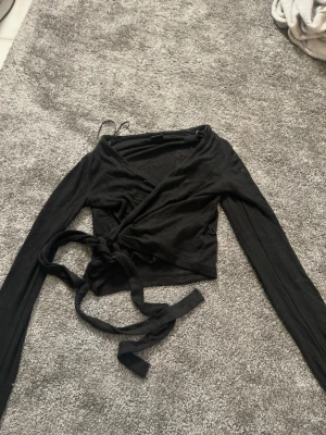 Snygg tröja från Gina  - Säljer en svart omlottopp från Gina Tricot i storlek XXS. Toppen har långa ärmar och knytband i midjan för en snygg passform. Materialet känns mjukt och stretchigt.