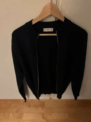 Svart ziptröja från MNG - Snygg svart ziptröja från MNG med ribbad krage och långa ärmar. Tröjan har en stilren design utan synliga loggor och är perfekt att slänga på sig över en t-shirt. Passar till både jeans och kjol för en clean look.