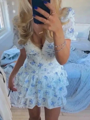 Populär miniklänning  - Söt kortklänning med blått blommönster. Klänningen har v-ringning, puffärmar och volangdetaljer i kjolen. Perfekt för sommarens alla tillfällen. Tillverkad i lätt och luftig polyester. Klänningen har en mindre fläck där bak