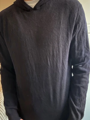 Svart hoodie från Oscar Jacobson - En enkel och stilren svart hoodie med huva och långa ärmar. Tillverkad i ett mjukt och bekvämt material som passar perfekt till en avslappnad look. Inga synliga loggor eller tryck för en clean stil. Den ser lite solblekt ut på bilden men det är skenet som bedrar. 