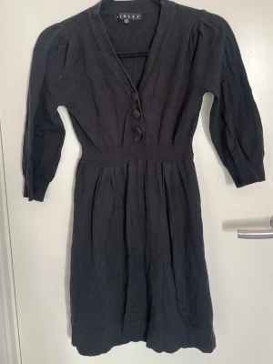 Svart stickad klänning från Sisley - Mysig svart stickad klänning från Sisley med v-ringning, tre dekorativa knappar framtill och trekvartsärm. Klänningen har markerad midja och lätt utställd kjol, perfekt för en stilren look.