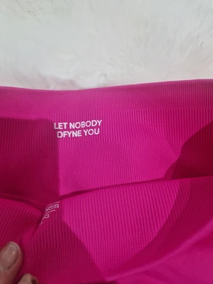 Rosa träningstights shorts från DFYNE - Snygga rosa träningstights shorts från DFYNE med hög midja och ribbad struktur. Perfekta för gymmet eller andra sportaktiviteter. Stretchigt material som ger en tight och bekväm passform. Logga på linningen och coolt citat på insidan.