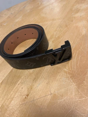 monogram skärp - Snyggt svart skärp från  med klassiskt monogrammönster och en stor  i metall som spänne. Bältet har en lyxig känsla med brunt innerfoder och passar perfekt till jeans eller kostymbyxor.