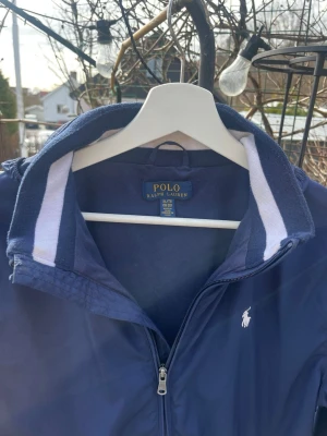 Marinblå vindjacka från Polo Ralph Lauren XS/S - Snygg marinblå vindjacka från Polo Ralph Lauren med dragkedja framtill, två fickor med dragkedja och klassisk vit logga på bröstet. Jackan har ståkrage med vit och blå detalj på insidan! Luvan går att ta av och jackan är i storlek XS/S        Riktig snygg nu till våren och sommaren som kommer☀️Nytt pris 1199kr mitt pris 299kr        Vid snabb affär går priset att diskuteras, frågor eller annat hör av dig, MVH Oliver 