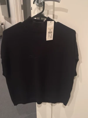 Svart stickad tröja XS - En svart stickad tröja med hög krage och korta ärmar. Tröjan har en diskret broderad logga framtill och ribbstickad kant vid halsen. Perfekt för dig som gillar en enkel och stilren look.