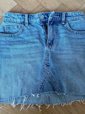 Hollister Jeansskjol - Säljer en snygg blå jeanskjol med fransig, rå kant nertill och klassiska fem fickor. Kjolen har bälteshällor, dragkedja och knapp framtill. Perfekt för dig som gillar en avslappnad och trendig stil.