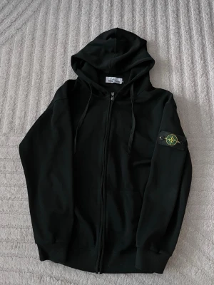 Svart hoodie från Stone Island - Snygg svart hoodie från Stone Island med dragkedja och klassisk logotyp-patch på vänster ärm. Hoodien har huva med dragsko och två fickor framtill. Perfekt för en stilren och avslappnad look.