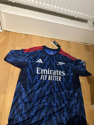 Arsenal blå matchtröja Adidas - Snygg blå Arsenal matchtröja från Adidas med blixtmönster och röda ränder på axlarna. Helt ny. Med tags på. Pris går att diskuteras. Öppen för frågor och bud. Mått kan skickas