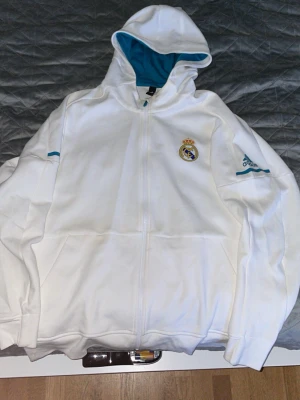 Vit Real Madrid hoodie från Adidas - Snygg vit hoodie från Adidas med Real Madrid-logga på bröstet och Adidas-logga på ärmen. Dragkedja framtill, huva med blått innerfoder och två fickor. Perfekt för dig som älskar fotboll och vill ha en sportig look. Tillverkad i mjukt material för extra komfort.