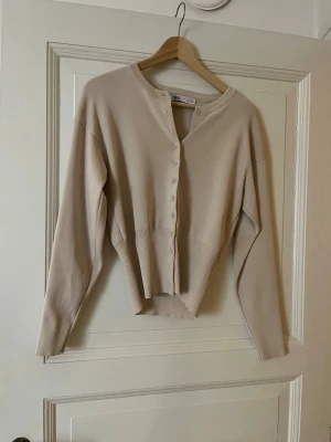 Beige ribbad kofta från Zara - Snygg beige kofta från Zara med ribbad midja och knappar framtill. Koftan har lång ärm och en croppad passform som ger en modern look. Perfekt att styla med både jeans och kjol.