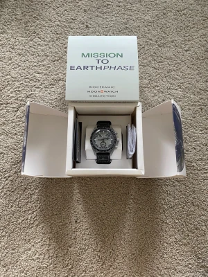 Omega x Swatch MoonSwatch Earthphase - Snygg Omega x Swatch MoonSwatch Mission To Earthphase med svart och grå urtavla, biokeramisk boett och svart band. Klockan har tachymeter och flera subdials, samt coola detaljer inspirerade av rymden. Perfekt för dig som gillar unika accessoarer.