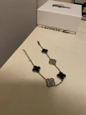 Silverfärgat armband med pärldetaljer - Snyggt armband i silverfärg med fyra fyrklöverformade hängen, två i svart och två täckta av små glittrande pärlor. Kedjan är tunn och elegant, perfekt för att ge en stilren touch till din outfit.